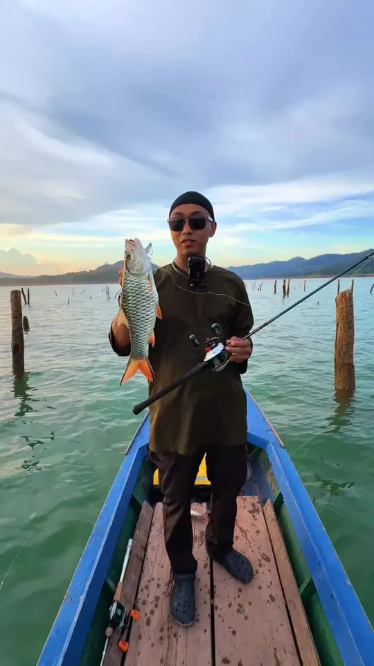 Abe Wea berdiri dalam perahu memegang ikan sebarau dan joran di tasik
