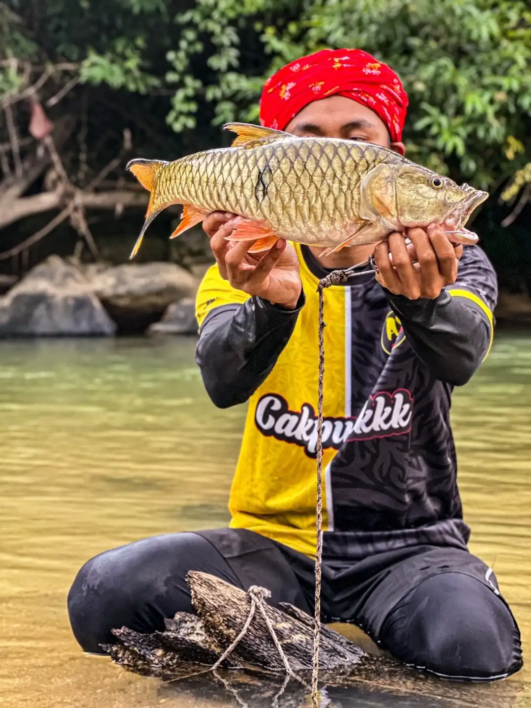 Abe Wea berkain semutar merah dan berjersi Cakpuakk, melutut di sungai sambil memegang ikan sungai