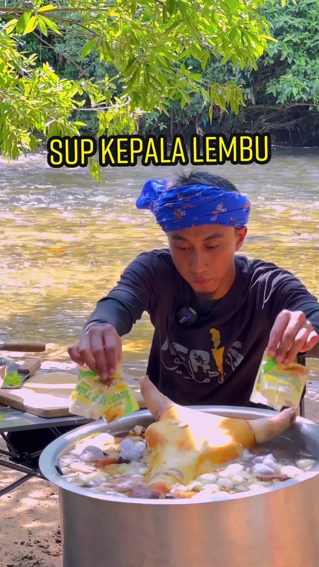 Abe Wea memasukkan rempah ke dalam periuk sup kepala lembu di tepi sungai, dengan tulisan SUP KEPALA LEMBU