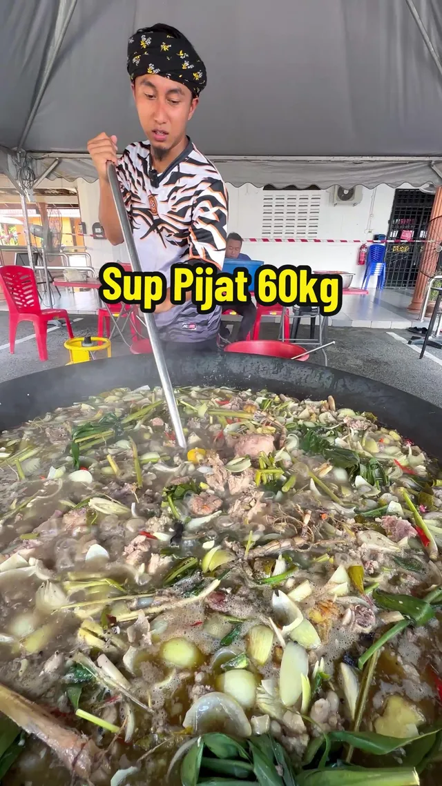 Abe Wea mengacau sup pijat dalam kawah besar di bawah khemah, dengan teks 'Sup Pijat 60kg'