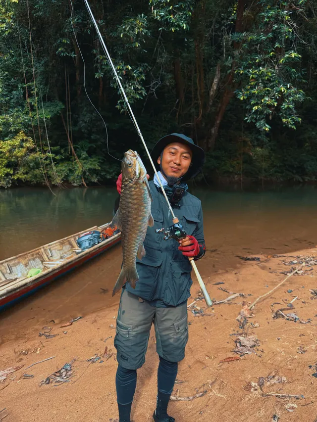 Abe Wea memegang ikan sungai dan joran di tebing berpasir Ulu Sungai Lebir, perahu di belakang
