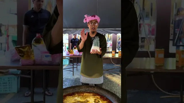 Abe Wea memegang plastik berisi bahan putih di belakang kawah gulai di bawah kanopi